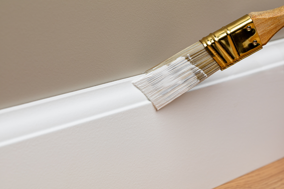 Someone applying white paint to a baseboard with a brush 
